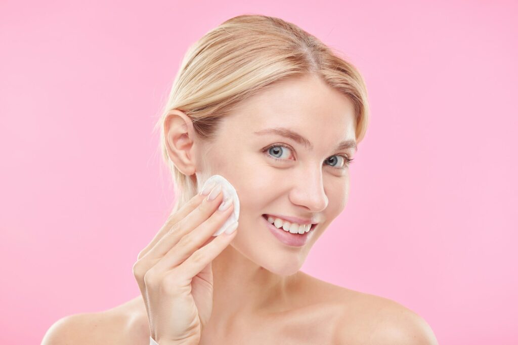 Pretty young smiling female with cotton pad applying toner or micellar water